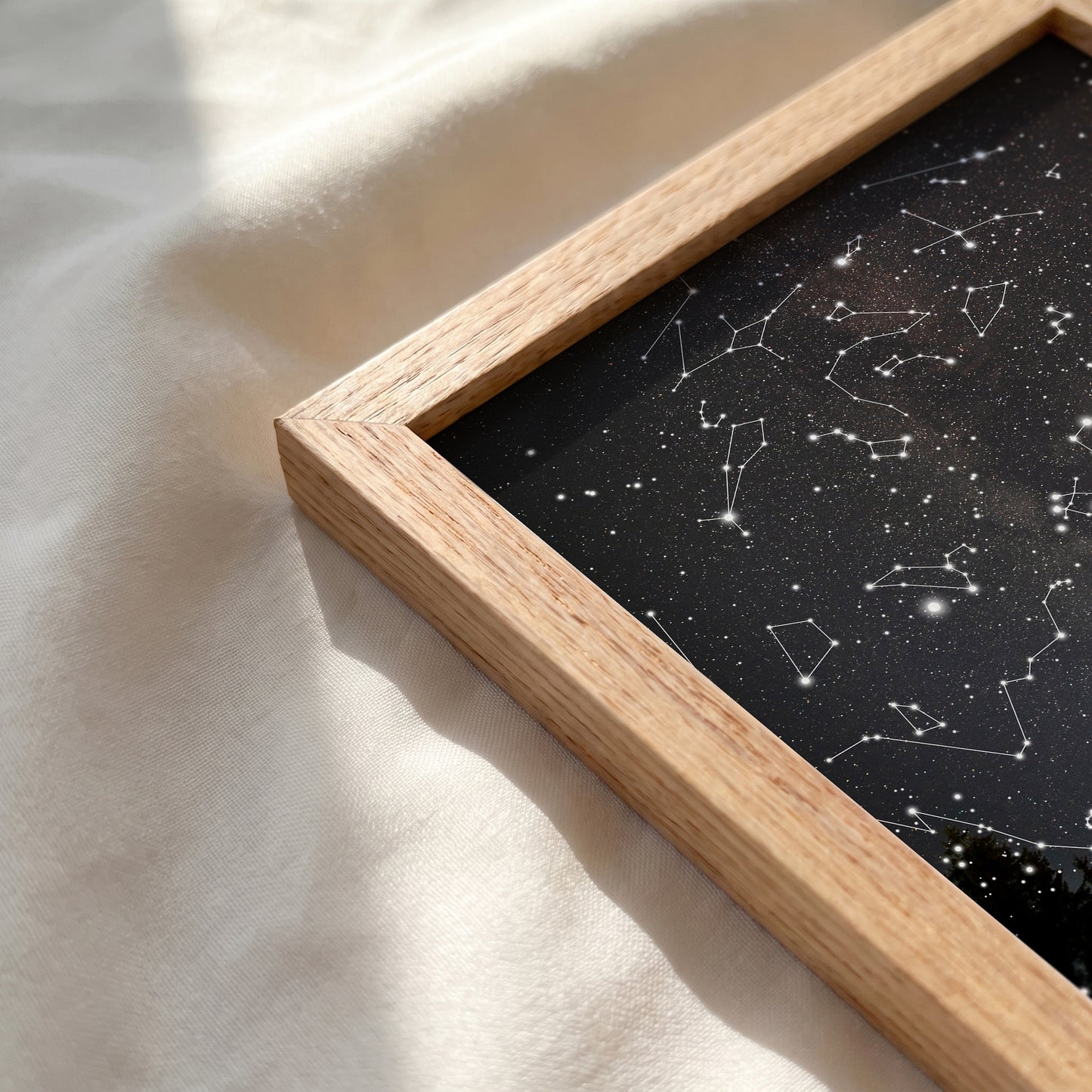 A close-up of a framed art print featuring a black starry sky with white constellations, set in a light natural wood frame and resting on a soft, sunlit cream fabric.