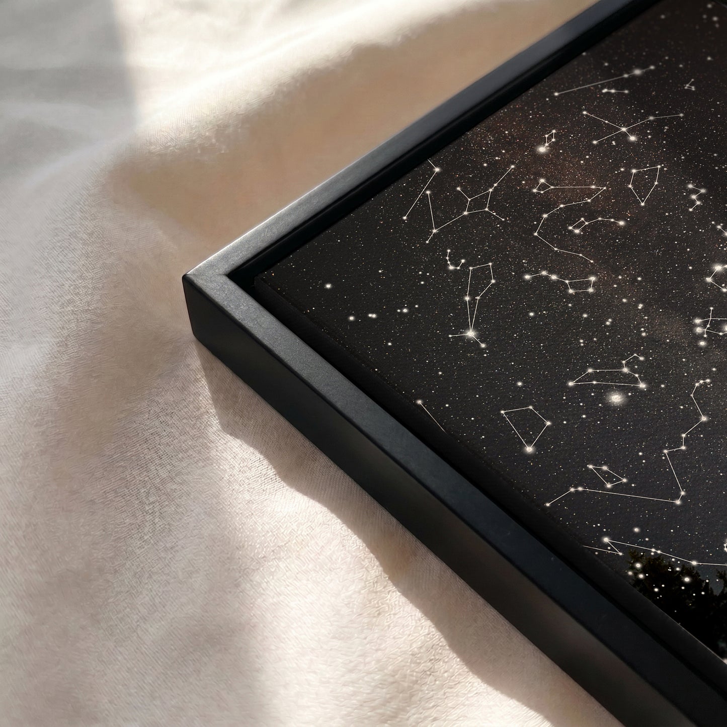 A close-up of a framed black art print featuring white constellation lines and stars, resting on a soft beige fabric surface with natural sunlight and shadows.