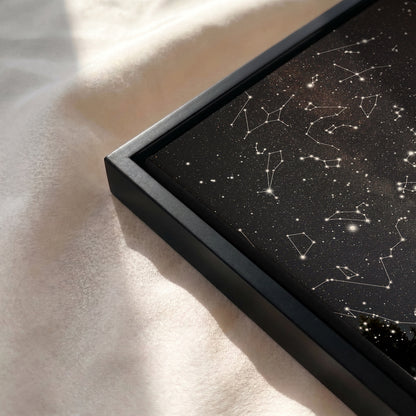A close-up of a framed black art print featuring white constellation lines and stars, resting on a soft beige fabric surface with natural sunlight and shadows.