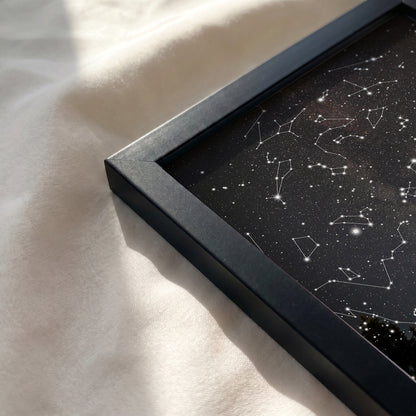 A close-up of a framed black starry sky art print with white constellation lines, resting on a soft white fabric surface with natural sunlight and shadows.