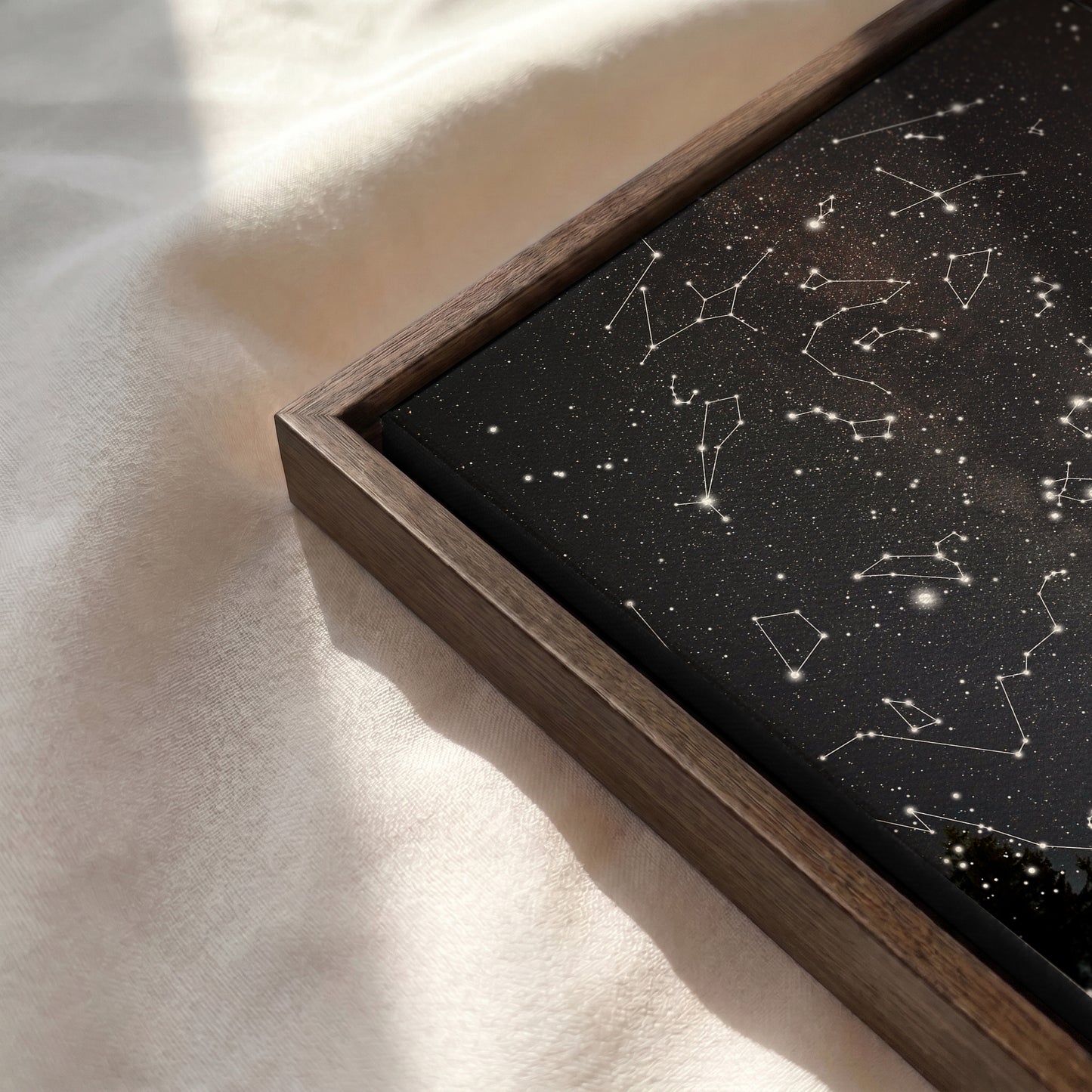 A framed art print featuring a black starry sky with white constellation lines, set in a dark wood frame and resting on a soft, sunlit cream fabric.