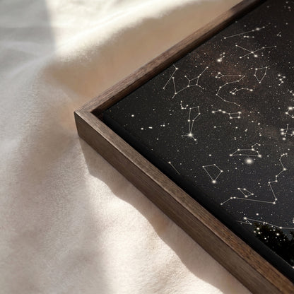 A framed art print featuring a black starry sky with white constellation lines, set in a dark wood frame and resting on a soft, sunlit cream fabric.