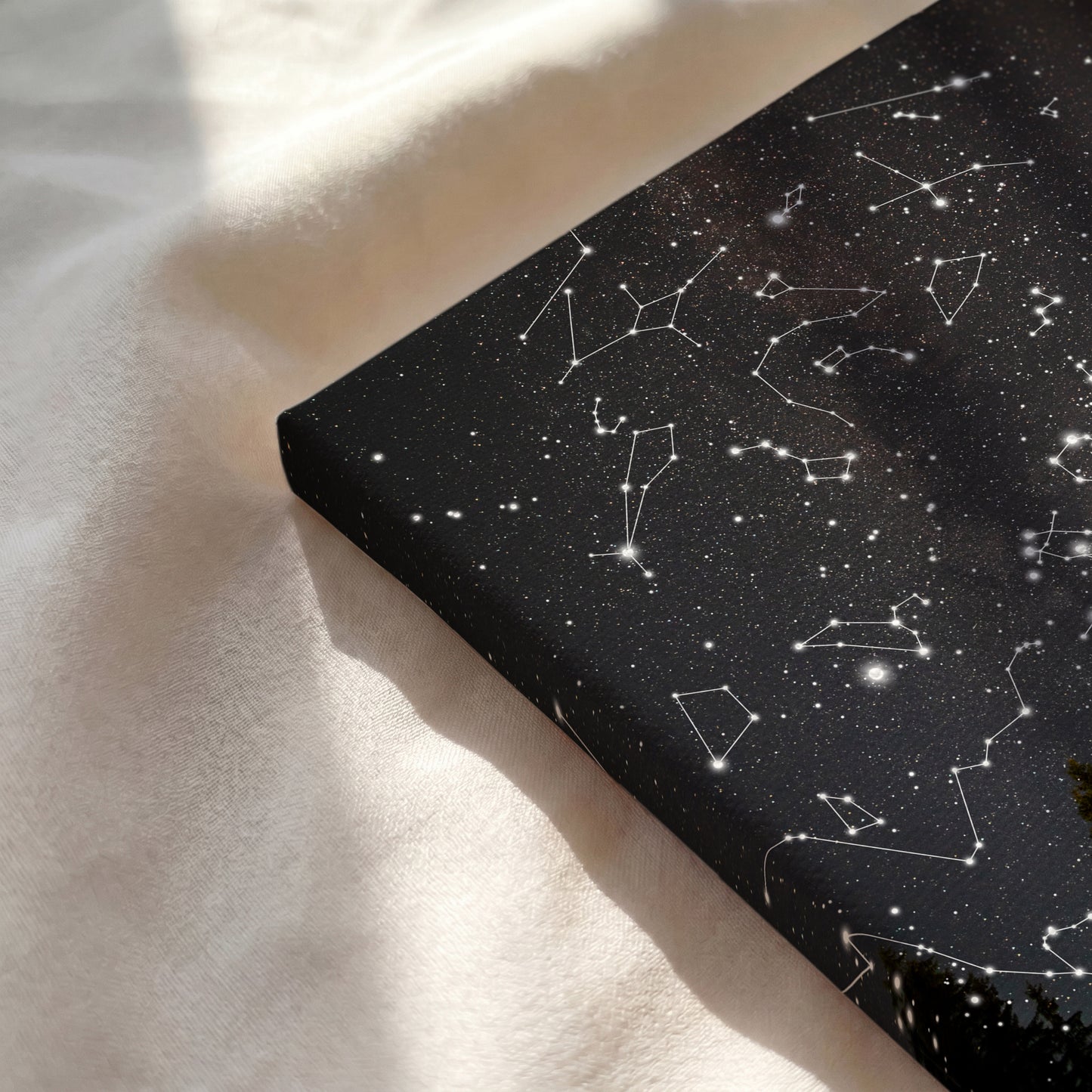 A rectangular black fabric or mat with a white constellations and star pattern, photographed on a soft cream fabric surface with natural sunlight and shadows.