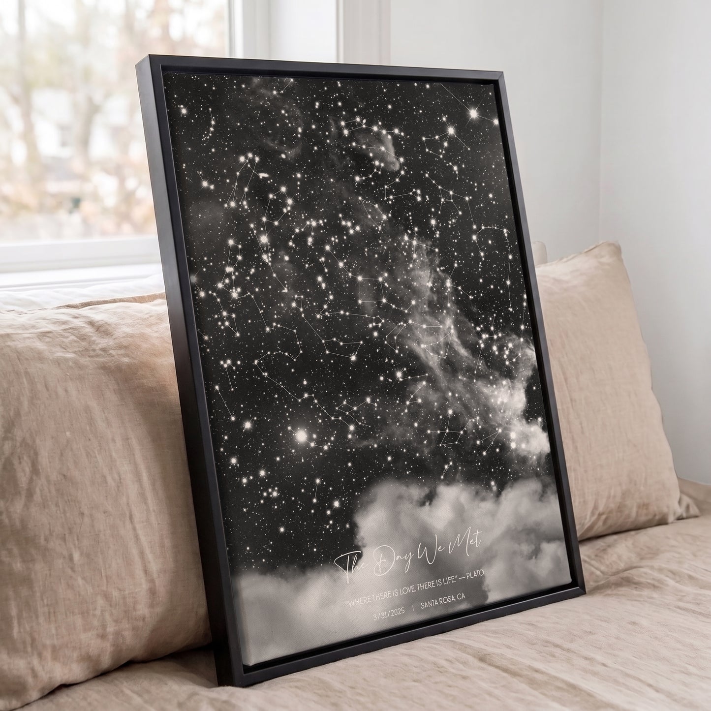 Framed personalized cloudy star map print displayed against beige pillows on a bed in a softly lit bedroom.