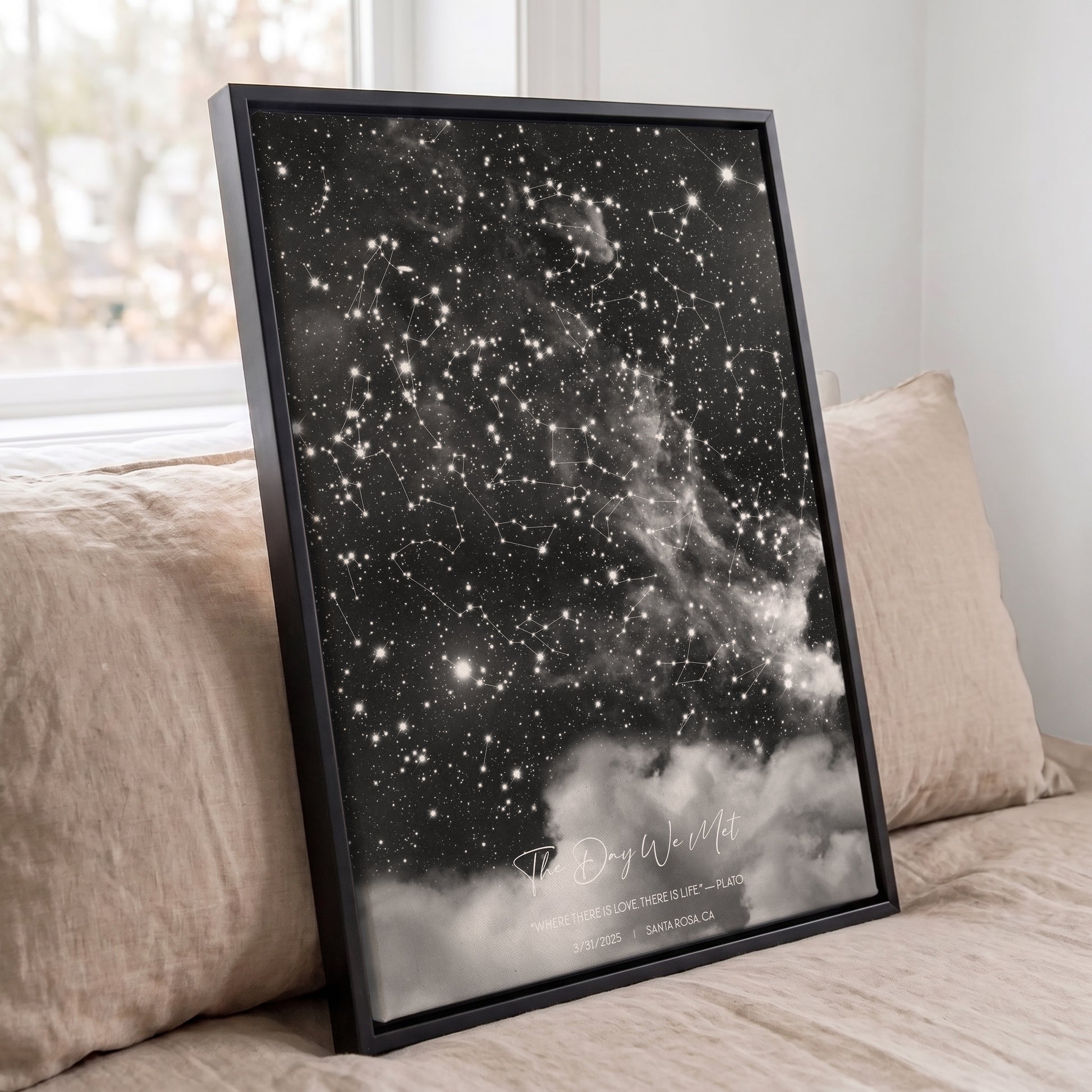 Framed personalized cloudy star map print displayed against beige pillows on a bed in a softly lit bedroom.