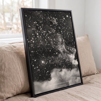 Framed personalized cloudy star map print displayed against beige pillows on a bed in a softly lit bedroom.