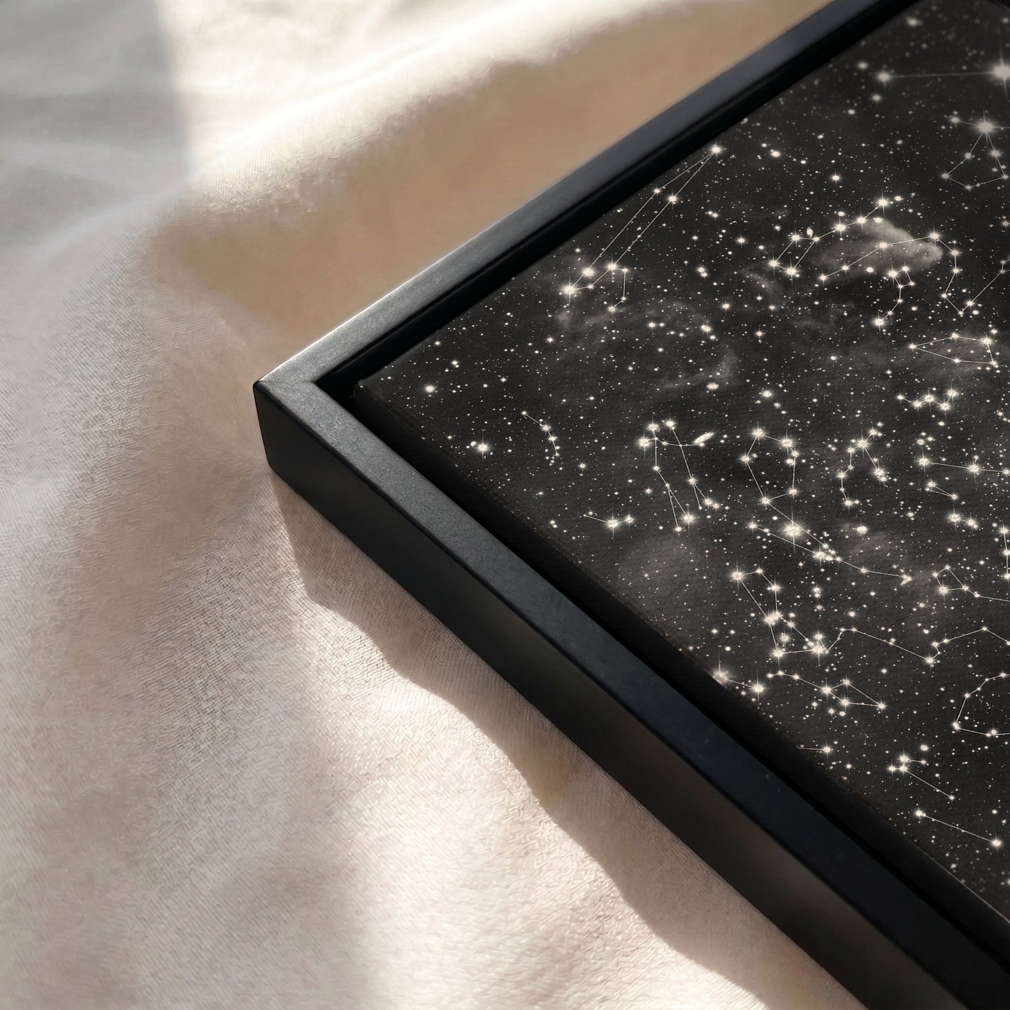 Close-up of a personalized cloud background star map canvas in a black wooden frame resting on a soft, sunlit cream-colored fabric surface.