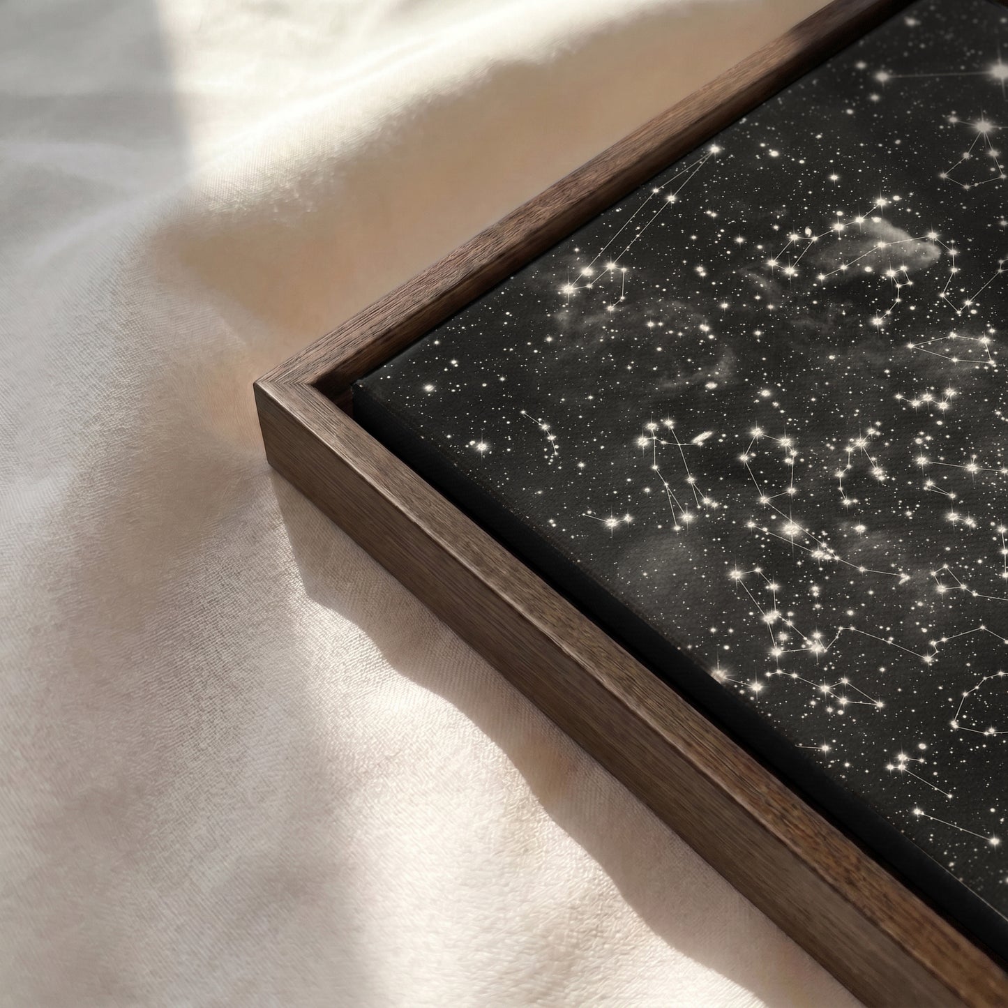 Close-up of a personalized cloud background star map canvas in a natural red oak frame resting on a soft, sunlit cream-colored fabric surface.