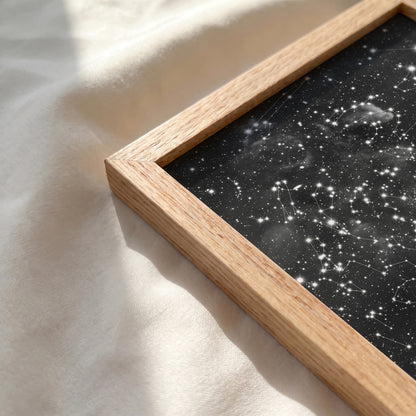 Close-up of a personalized cloud background star map print in a natural red oak frame resting on a soft, sunlit cream-colored fabric surface.