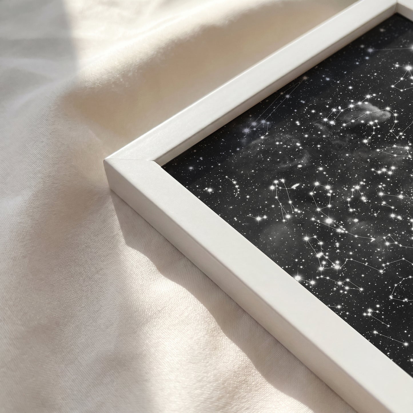 Close-up of a personalized cloud background star map print in a white wooden frame resting on a soft, sunlit cream-colored fabric surface.