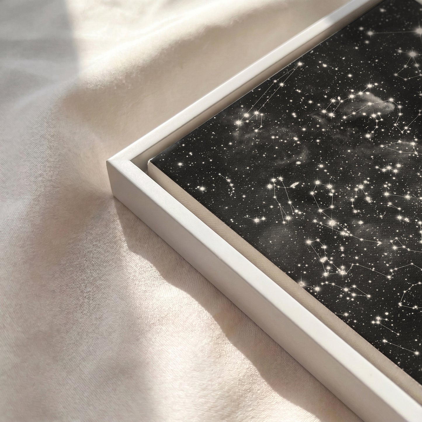 Close-up of a personalized cloud background star map canvas in a white wooden frame resting on a soft, sunlit cream-colored fabric surface.