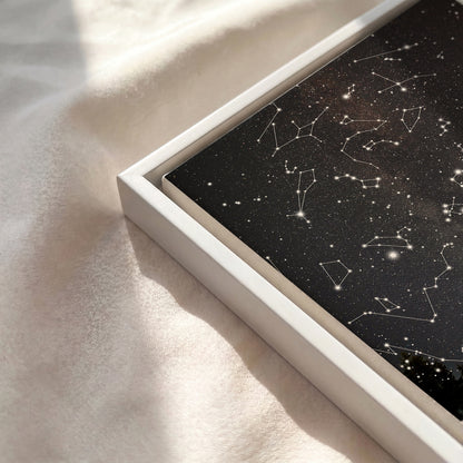A framed art print featuring a black starry sky with white constellations, set in a white frame and resting on a textured cream fabric under soft sunlight.