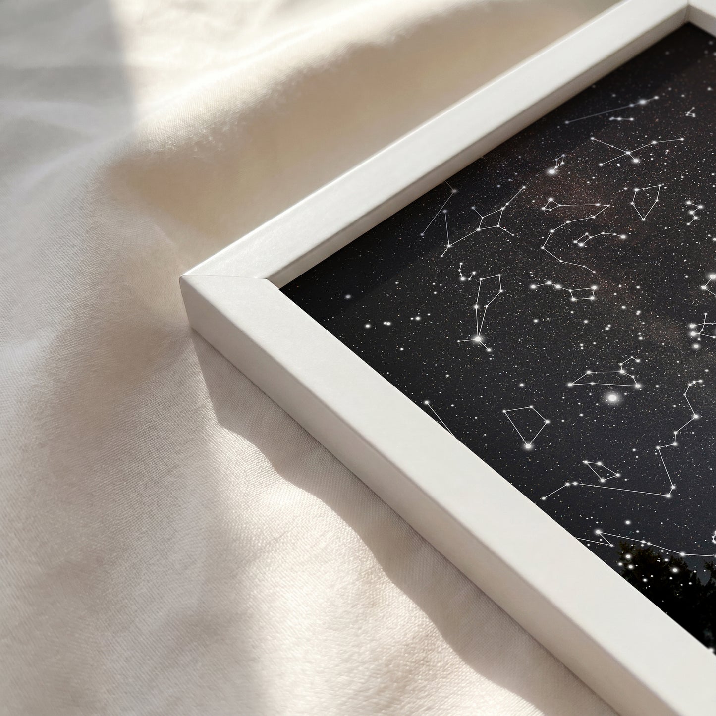 A framed black starry sky art print with white constellation lines, resting on a soft beige fabric surface with warm sunlight and shadows.